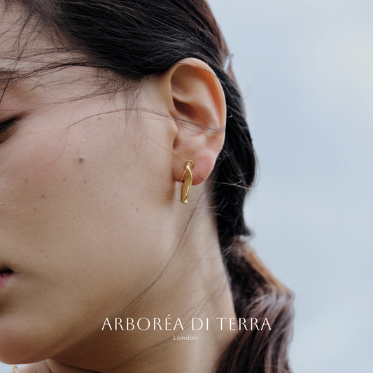 Gold earring worn by a person with a blurred background, featuring the brand 'Arboréa di Terra'.