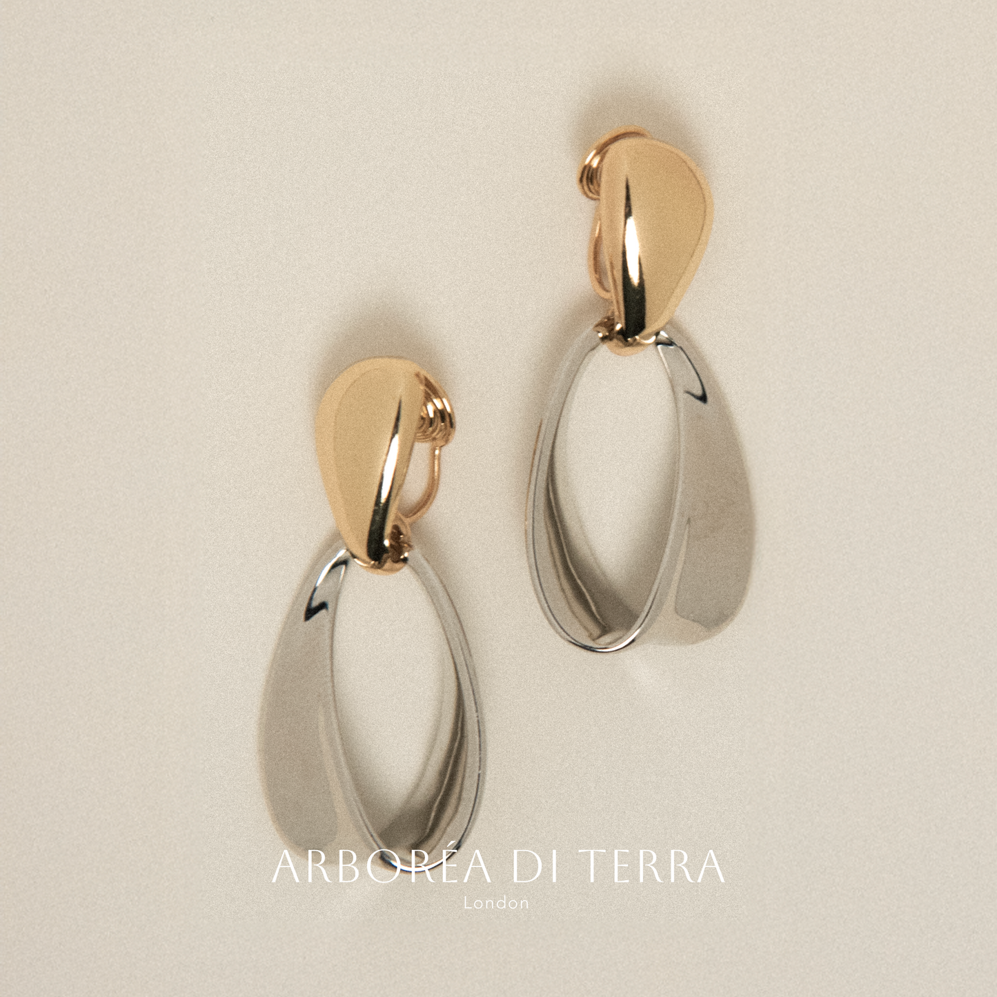 Pair of gold and silver earrings on a beige background with 'Arborea Di Terra' branding.
