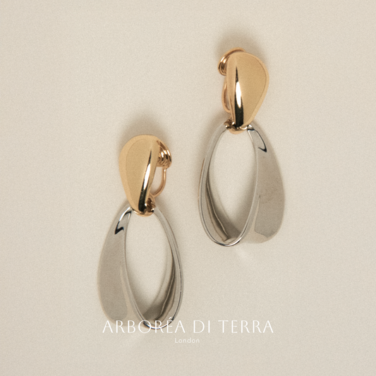 Pair of gold and silver earrings on a beige background with 'Arborea Di Terra' branding.
