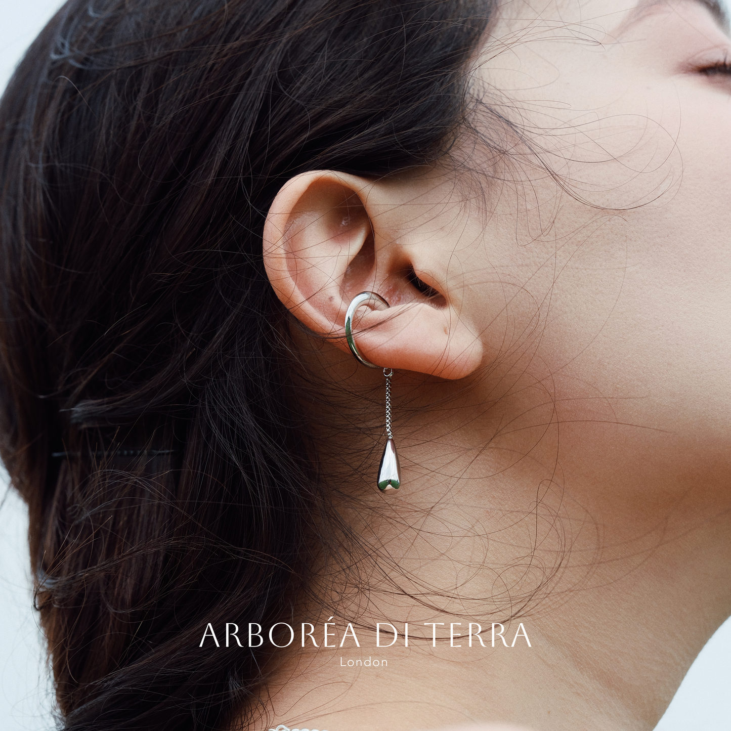 Close-up of an ear wearing a silver earring with 'Arboréa di Terra' branding.
